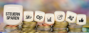 Holzwürfel mit der Aufschrift „Steuern sparen“ sowie Symbolen für Geld, Wachstum und Finanzplanung stehen auf gestapelten Münzen und veranschaulichen das Thema Steuern sparen als Selbstständiger & Freiberufler
