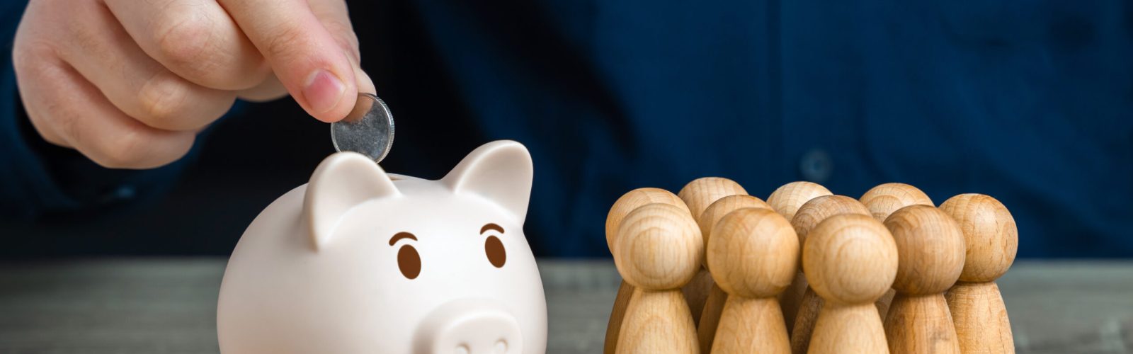 Hand wirft Münze in ein Sparschwein neben Holzfiguren – Symbol für Vermögenssicherung, Gemeinschaft und steuerliche Optimierung.