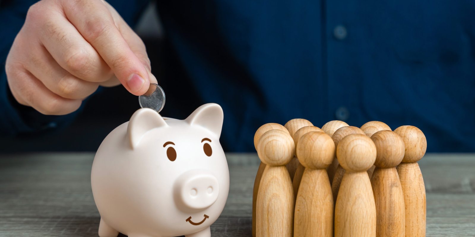 Hand wirft Münze in ein Sparschwein neben Holzfiguren – Symbol für Vermögenssicherung, Gemeinschaft und steuerliche Optimierung.